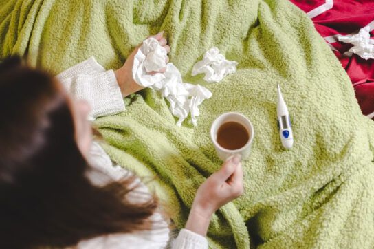 A person with a cold, the flu, or a sinus infection surrounded by tissues and a thermometer holding a cup of tea.