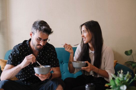 Adult male and adult female sitting on a sofa together each with a bowl of food, smiling and enjoying each others company possibly wondering how long does it take for Cialis to work