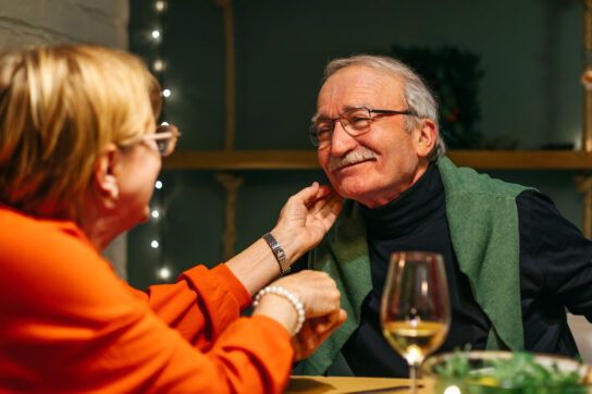 Older adult couple having dinner together, the male smiling as the female gently touches his face as they may wonder how to get viagra