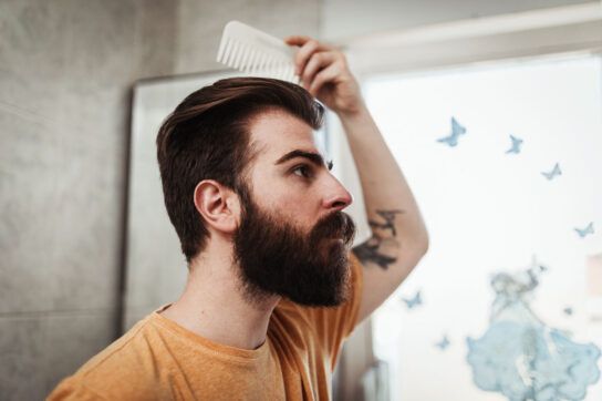 Side view of an adult male's head and shoulders who is combing their hair and appears to be looking in a mirror possibly wondering does testosterone cause hair loss?
