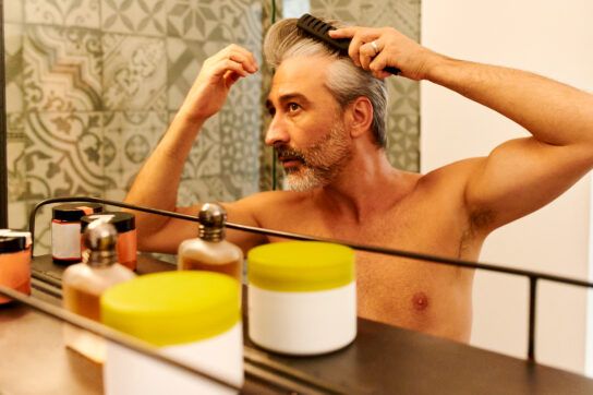 An older adult combing his hair, looking in the mirror, wondering how long minoxidil takes to work.