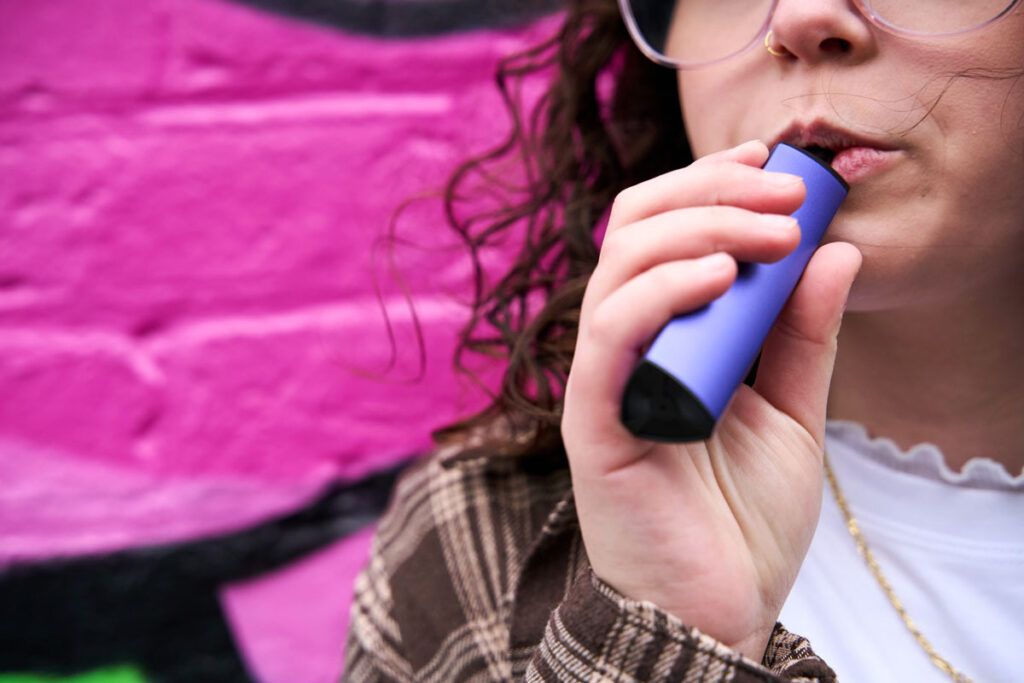 A young adult, shown from the nose down, vaping against a pink background, which may lead to or worsen COPD.