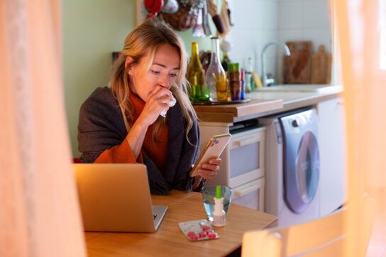 Adult female sitting at a kitchen table with a laptop and her phone in her left hand. Holding a tissue to her mouth to trap a cough, she could be looking for cough medicine for high blood pressure