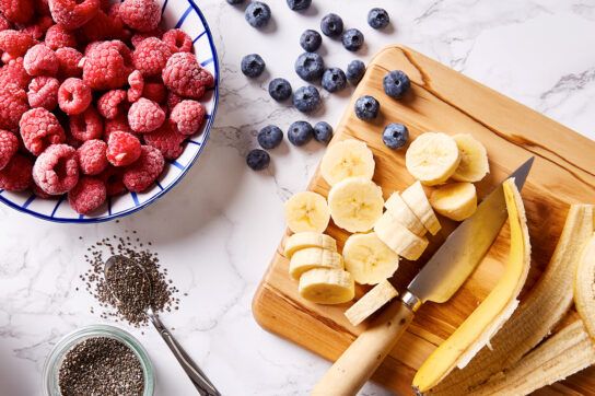 Overhead view of kitchen counter with a bowl containing raspberries on the left, blueberries scattered to the right, a chopping board with chopped banana on the left and chia seeds at the bottom in a small bowl and overflowing from a spoon representing foods you may find when researching how to lower your cholesterol