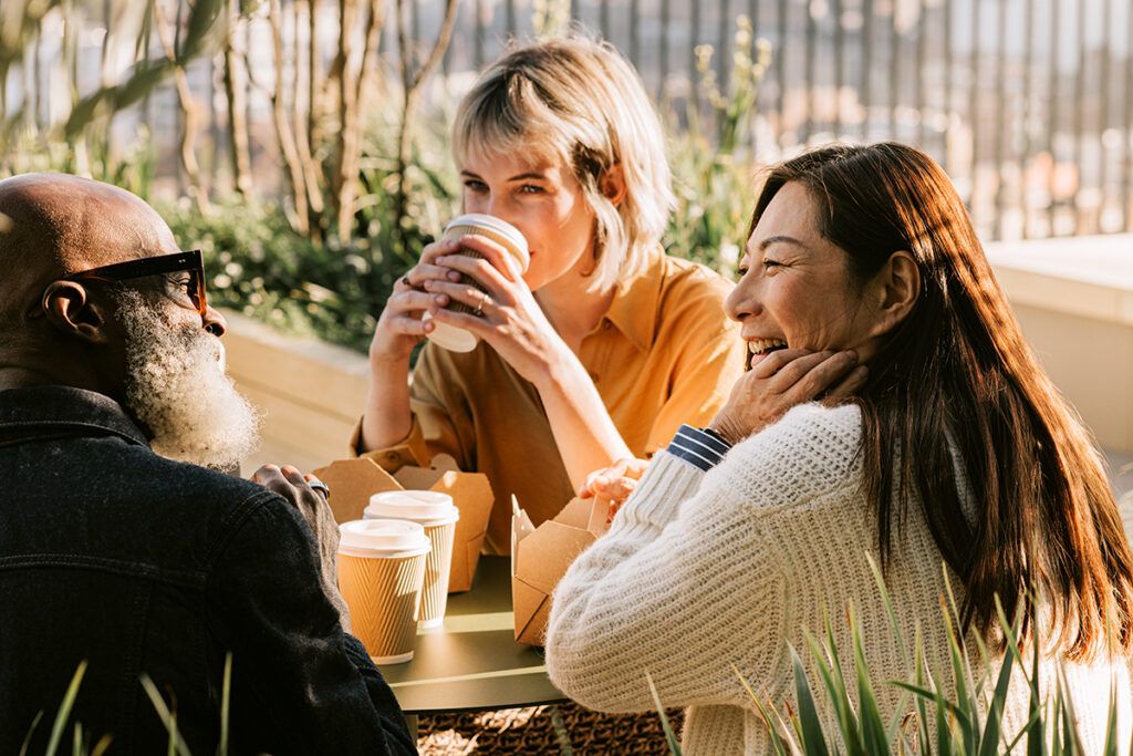 A group of three friends eating and laughing together outside over coffee.