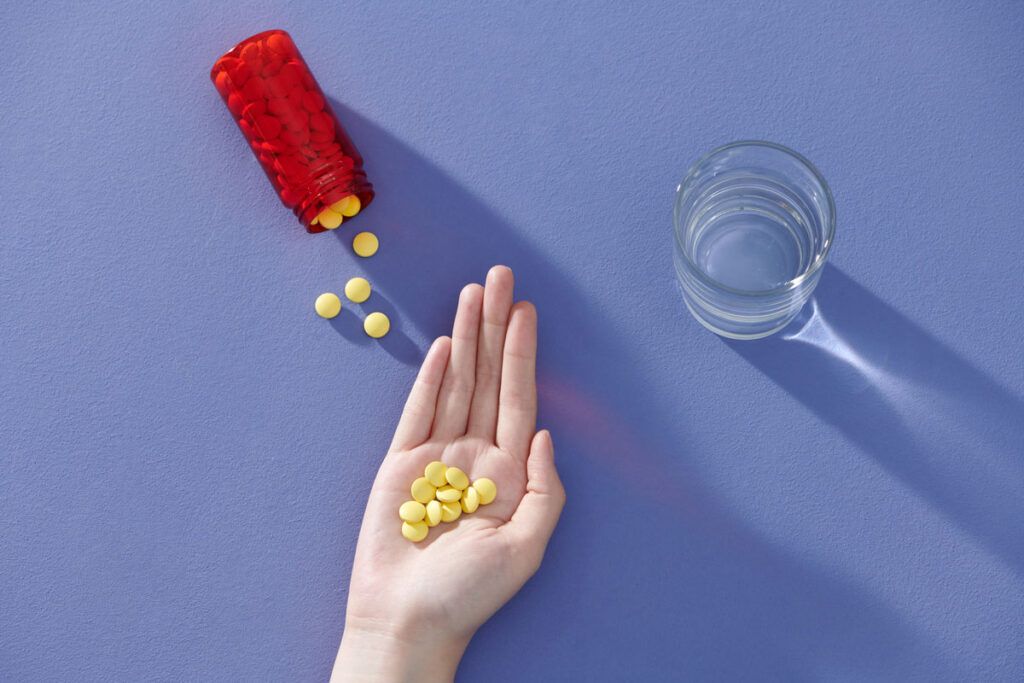 A bottle of vitamin E pills for cholesterol spilling onto a table and a hand holding some of the yellow pills next to a glass of water..