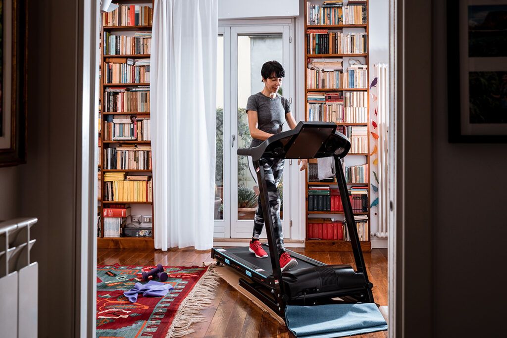 Older female walking on a treadmill with bookshelves on the wall behind her.