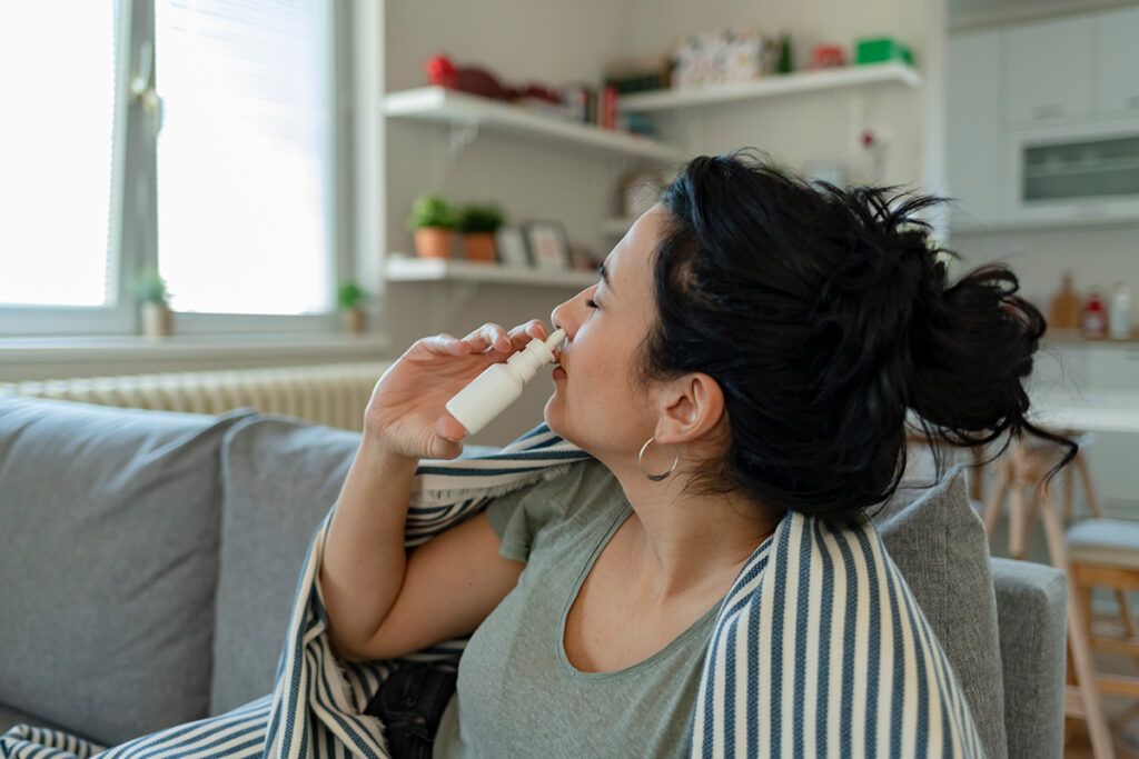 Adult female half laying on a sofa using a bottle of nasal spray correctly to stop the nasal spray going down their throat