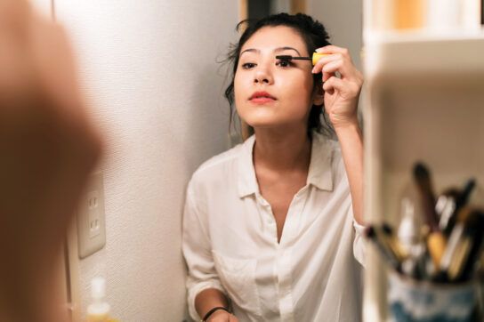 An adult applying mascara while looking in a mirror.