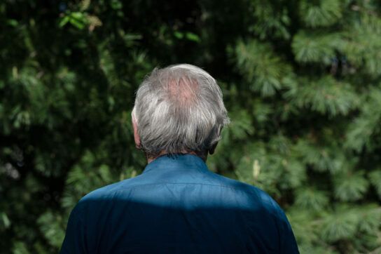 View of a persons head and shoulders from behind, standing in front of some evergreen trees, possibly wondering the best time to take finasteride for hair loss