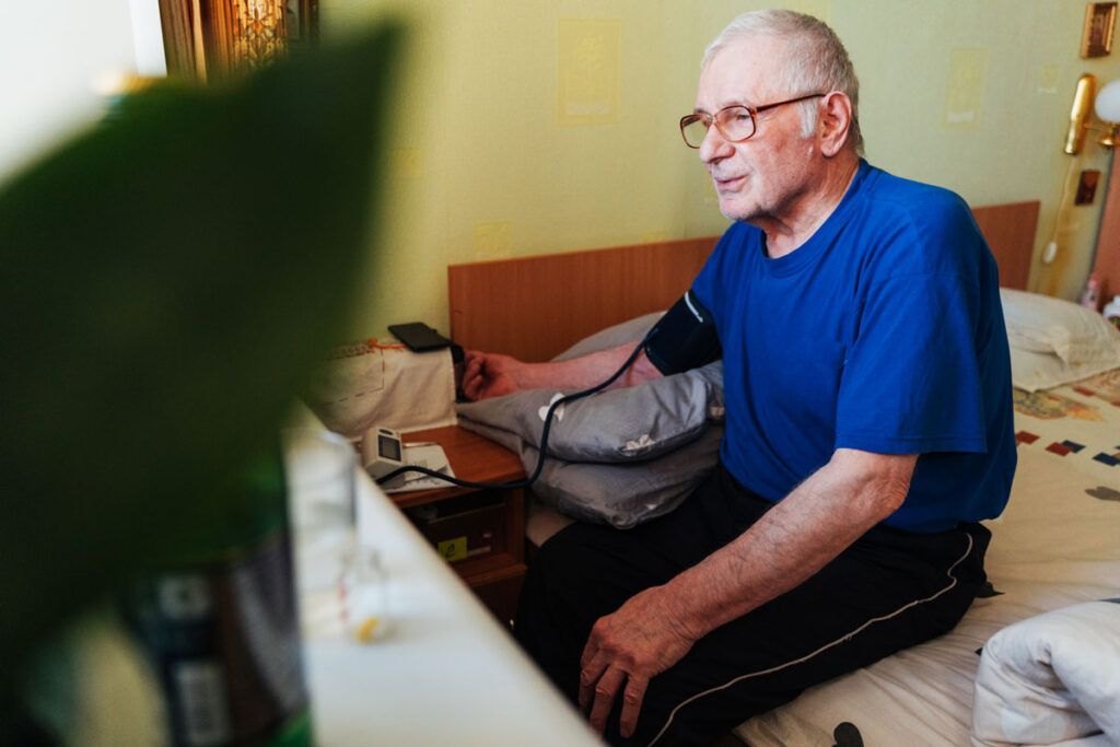 Older adult sat on a bed checking his blood pressure via an at home monitor.