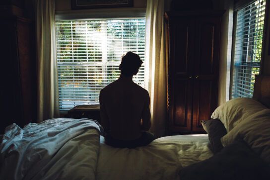 The back of an adult male sitting on the side of a bed looking out of the window that has morning sunlight shining through the slats of a blind, possibly thinking can stress cause ED