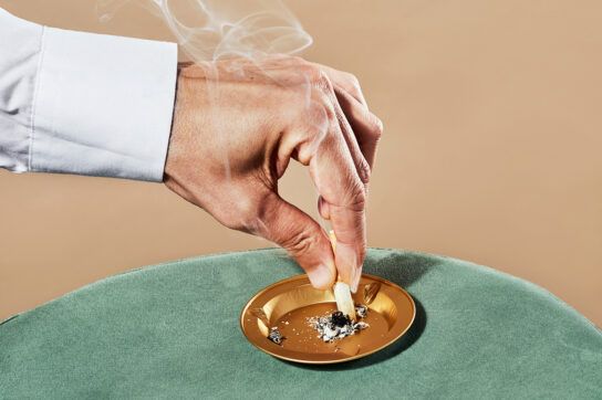 Close-up of a person’s hand putting a cigarette out on a gold ash tray.