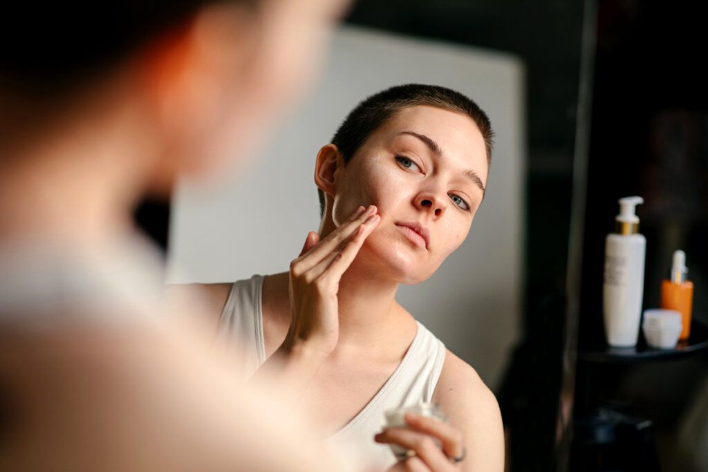 A young adult applying cream to their face in a mirror.