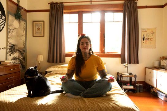 Adult female sitting on a bed cross legged in a meditative position with a shorthaired cat to her left possibly trying to overcome anxiety physical symptoms