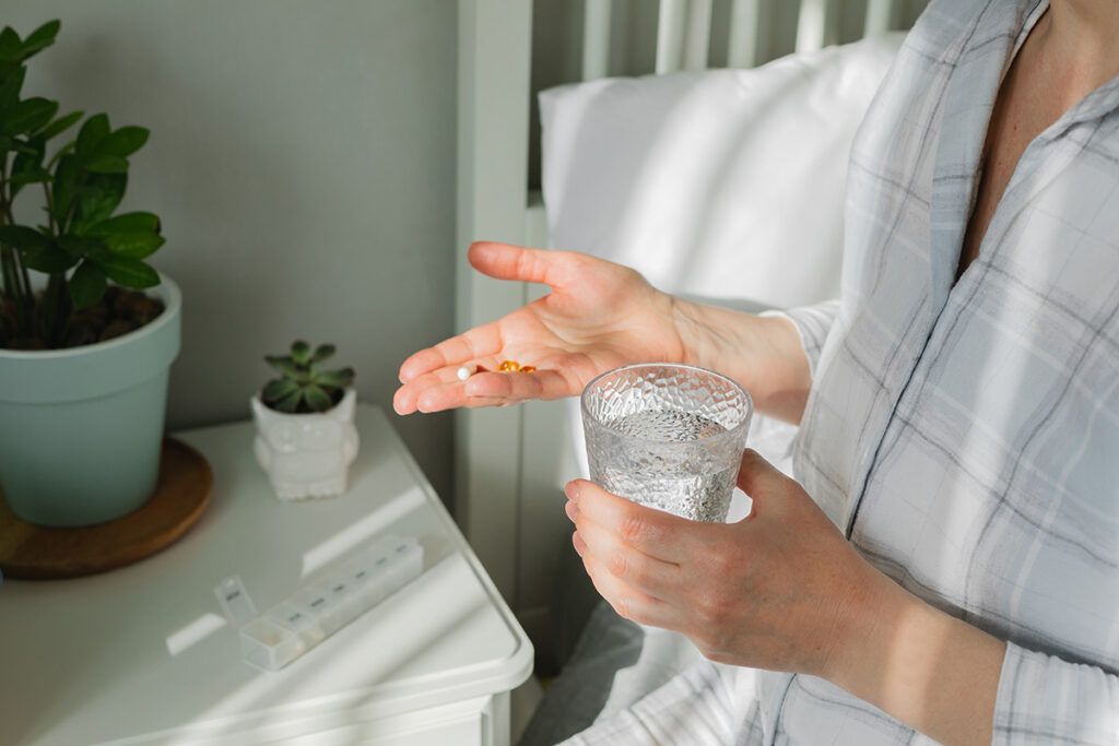 Woman sitting on side of bed holding pill in one hand and glass of water in other
