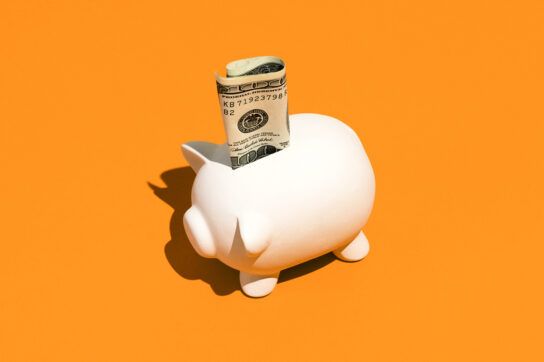 A piggy bank on an orange background with some folded dollars sticking out of the top.