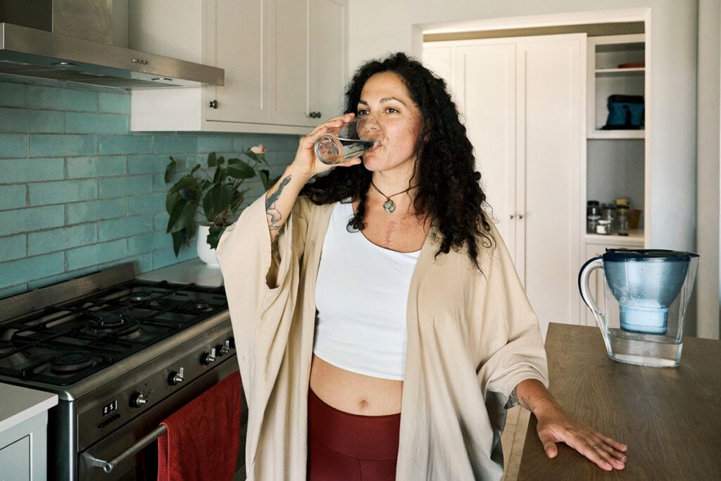 Person standing in a kitchen drinking a glass of water to depict a dry mouth.
