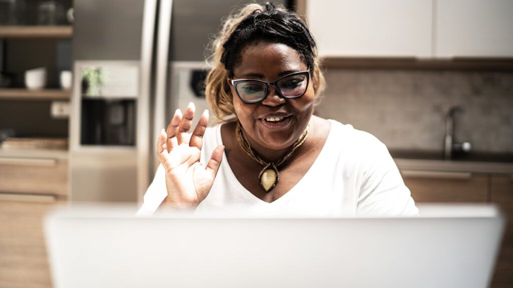 A woman on a video call with an online doctor, waving at her laptop.