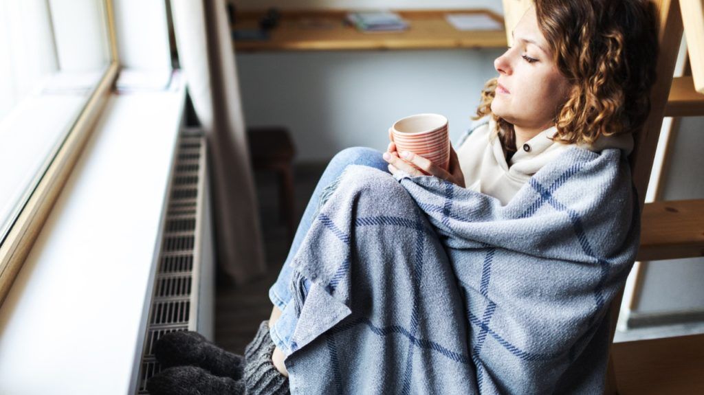A young woman in a blanket drinking tea, looking out of the window.